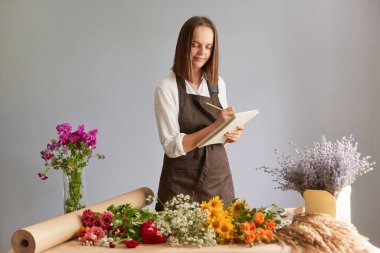 Çiçekçi dükkanında çiçek buketi yapan sevimli bir kadın. Gri arka planda tek başına duruyor. Not defterine sipariş yazıyor..