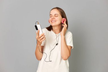 Beyaz tişörtlü, çiçekli kulaklık takan ve akıllı telefonlu müzik dinleyen çekici esmer kadın gri arka planda gözlerden uzak duruyor..