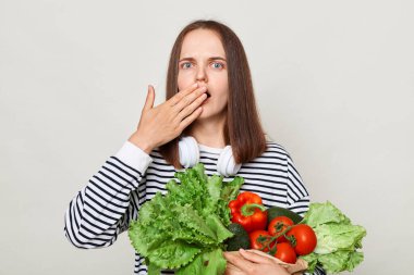 Beslenme bakımı ve seçenekler. Vejetaryen dostu yemek seçenekleri. Market alışverişi. Gri arka planda izole edilmiş sebze taşıyan çizgili bir tişört giyen şaşkın kadın.