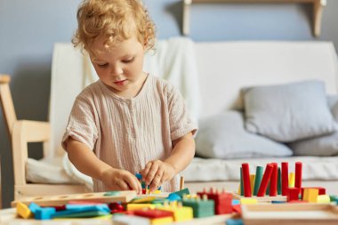 Anaokulu aktiviteleri. Renkli eğitici oyunlar. Küçük çocuğun büyük merakı. Konsantre dalgalı saçlı küçük sarışın bebek ev içinde ahşap eğitici oyuncaklar oynuyor..