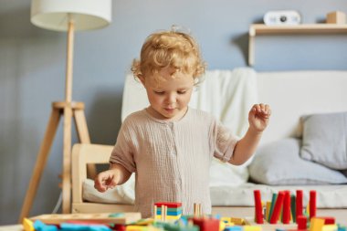 Neşeli bir anaokulu çocuğu yapmak. Bebek yuvası maceraları işte. Oynayarak öğreniyorum. Odaklanmış dalgalı saçlı sarışın bebek evde ahşap eğitici oyuncaklar oynuyor..