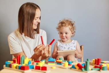 Meraklı çocuğun eğlenceli öğrenmesi. Eğlence için tahta oyuncaklar. Çocuk odasında birlikte keşfe çıkmıştık. Gülümseyen anne ve kız, gri arka planda saklanmış tahta oyuncaklarla oyun oynuyorlar.