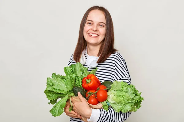 采购杂货 享受绿色饮食 新鲜蔬菜和蔬菜 身穿条纹休闲衫 背负着灰色背景蔬菜的微笑女人 — 图库照片
