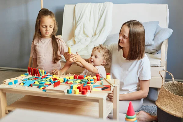 Babysitter\'s creative playtime. Learning through play and toys. Motherhood and family relationships. Smiling mother playing with little daughters engaged in game activity at home interior