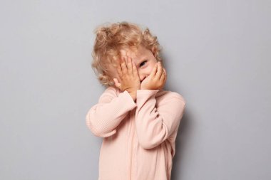 Sevimli bebek portresi. Sarı dalgalı saçlı, gri arka planda izole edilmiş gül rengi tişörtlü, utangaç bir kız bebek yüzünü kollarıyla kameraya doğru bakıyor.