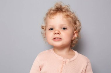 Gri arka planda izole edilmiş sarı dalgalı saçlı, neşeli, kendine güvenen bebek portresi gururlu bir ifadeyle kameraya bakıyor.