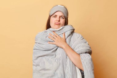 Mutsuz, uykulu, uyku maskesi takmış, yorgana sarılmış, bej arka planda izole edilmiş, dinlenen ve somurtkan suratıyla şekerleme yapan mutsuz, uykulu kadın.