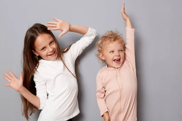 非常に幸せな小さな女の子姉妹は一緒にダンスを楽しみながら 灰色の背景に隔離された陽気な喜びの表情で笑いながら腕を上げました — ストック写真