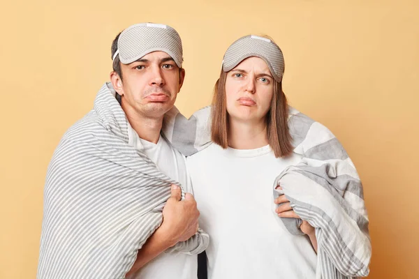 Sad unhappy couple man and woman wrapped in blanket isolated over beige background looking at camera with sorrow want to sleep more early wake up.