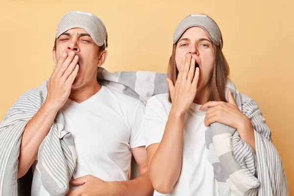 Sleepless couple man and woman wrapped in blanket isolated over beige background needs rest covering mouth with palm yawning needs rest having nap