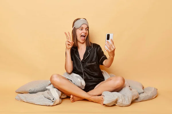 Cheerful woman sitting on floor wrapped in plaid wearing pajama, sleep eye mask isolated over beige background having video call on smartphone showing v sign.