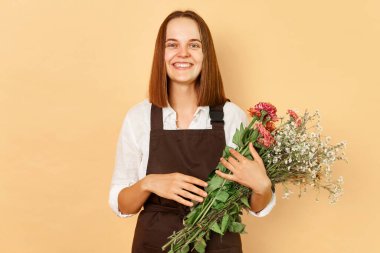 Çiçek tasarımcısı. Bitki dükkanı. Taze çiçekler. Botanik güzellik. Kahverengi önlük giyen, bej arka planda saklanmış çiçekleri tutan, gülümseyen beyaz bir kadın.
