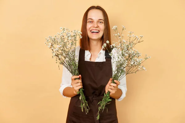 Floral arrangement. Professional florist. Botanical beauty. Cheerful joyful young caucasian woman florist wearing brown apron holding flowers isolated over beige background