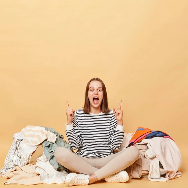 Heyecanlı beyaz kadın, bej arka planda izole edilmiş rengarenk kıyafetlerin yanında poz veriyor. Reklam alanına işaret ediyor..