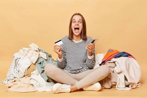 Aşırı neşeli Kafkasyalı kadın, bej arka planda izole edilmiş, rengarenk kıyafetlerin yanında poz veriyor. Elinde cep telefonu ve kredi kartı ile sevinçten havalara uçuyor..