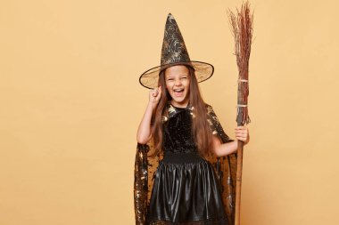 Ürkütücü büyücülük geleneği. Sihirli büyücü ve büyücü. Uzun saçlı, siyah pelerinli, bej arka planda izole edilmiş süpürgesi olan çılgın heyecanlı küçük kız.