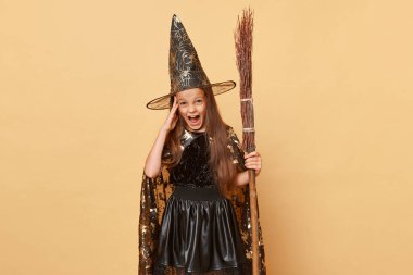 Ekim gizem karnavalı. Uzun şapkalı kötü cadı. Uzun saçlı küçük kız, cadılar bayramını kutlayan bej arkaplan çocuğu üzerinde izole edilmiş süpürge taşıyan siyah cadı kostümü ve karnaval şapkası giyiyor.