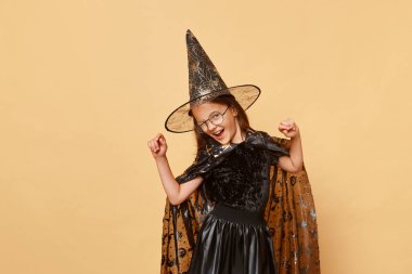 Son derece mutlu, neşeli, cadı kostümü giyen küçük bir kız. Panayır şapkası ve gözlük takıyor. Bej arka planda izole edilmiş..