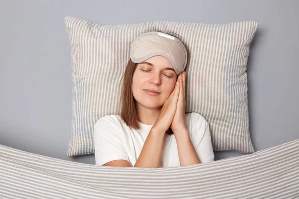 Calm relaxed young woman wearing white T-shirt and sleep eye mask lies under blanket in bedroom isolated on gray background leaning on her palms closed eyes having nap.