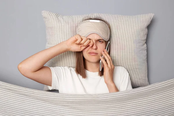 Sleepy young woman wearing white T-shirt and sleep eye mask lies under blanket in bedroom isolated on gray background talking on smartphone feeling sleepy rubbing her eye.