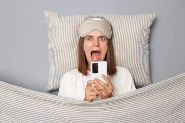 Shocked scared young woman wearing white T-shirt and sleep eye mask lies under blanket in bedroom isolated on gray background screaming with open mouth browsing internet with fear on face