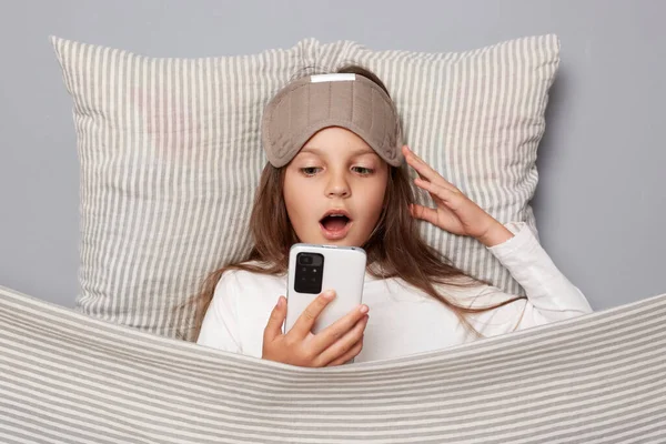 Shocked little girl wearing white shirt and sleep eye mask lies under blanket in bedroom isolated on gray background holding cell phone reading news looking at display with open mouth