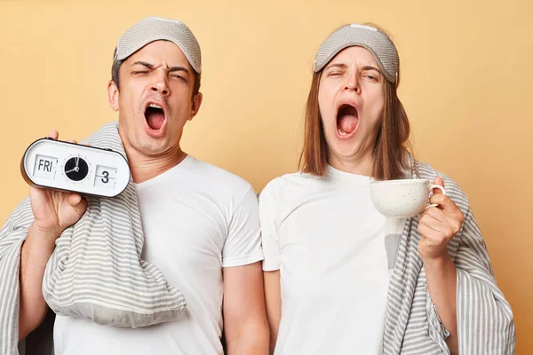 Sleepy couple man and woman in sleep eye mask wrapped in blanket holding coffee cup and alarm isolated over beige background yawning needs rest.