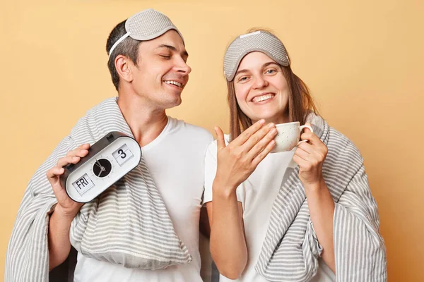 Smiling couple man and woman in sleep eye mask wrapped in blanket holding coffee cup and alarm isolated over beige background waking up in good mood enjoying hot beverage.