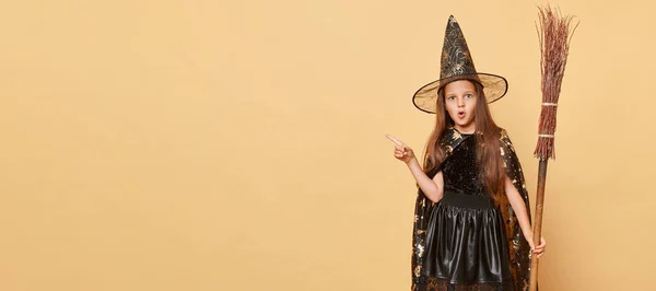 魔法に魅了された子供時代 プロモーションのためのコピースペースを指すベージュの背景の子供の上に隔離されたブロムの長い髪の黒いクロークの服を持つ驚いた少女 — ストック写真
