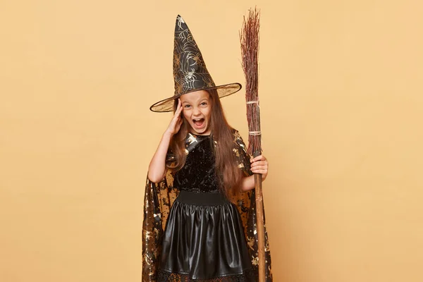 10月のミステリーカーニバル 背の高い帽子に魔女がいる 長い髪の小さな少女は ハロウィーンを祝うベージュの背景に隔離されたブルームを保持する黒い魔女の衣装とカーニバル帽子コーン — ストック写真