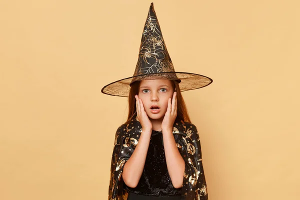 魔法の魔法使いと魔法使い ベージュの背景に隔離された魔女の衣装とカーニバルの円錐形の帽子を身に着けている驚くべき少女は恐ろしい何かで大きな目で見て頬に手を保ちます — ストック写真