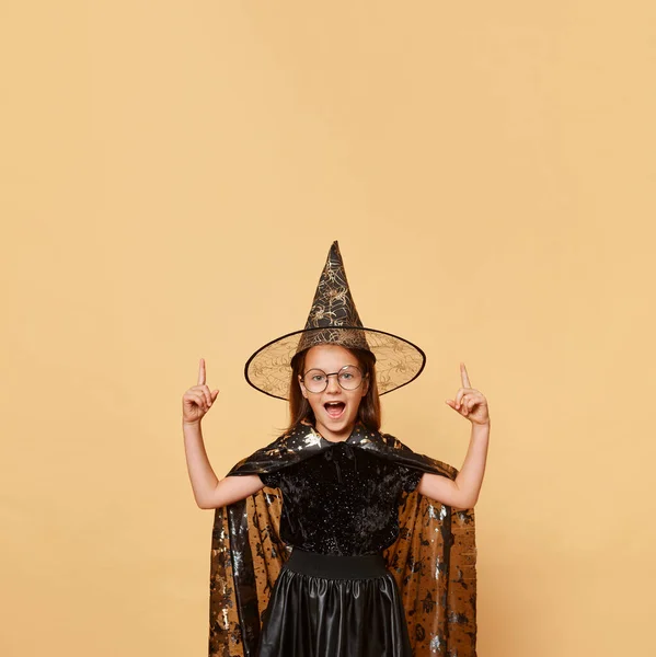 魔女のコスチュームカーニバルコーンハットを着てエキサイティングなクレイジーな少女は 広告エリアを指し示す驚きとベージュの背景の叫びの上に隔離された眼鏡 — ストック写真