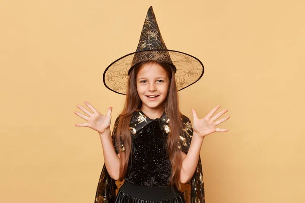 かわいい少女魔女の魔女の魔女の衣装を着て カーニバルコーン帽子はベージュの背景に隔離され ハッピーフェイスでハロウィーンの魔法の呪文を作る彼女の腕を上げました — ストック写真