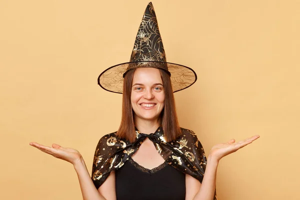 笑顔の若い女性は魔女のコスチュームとカーニバルコーンハットを着て ハロウィーンを祝い ベージュの背景に隔離され プロモーションのためのコピースペースを提示する手のひらを広げます — ストック写真