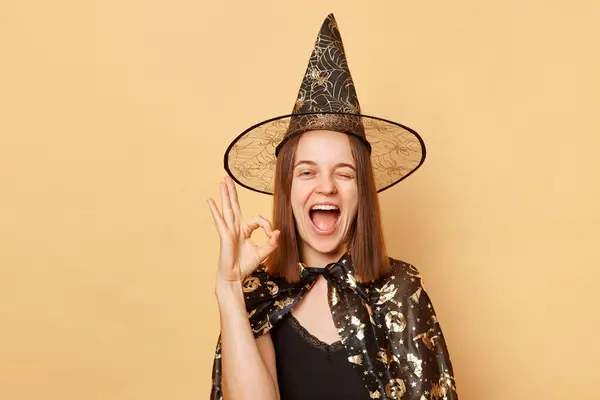 笑顔の若い女性は魔女の衣装を着て ハロウィーンを祝うカーニバルコーンハットは 大丈夫なジェスチャーを示すカメラにベージュの背景ウィンキングの上に隔離されました — ストック写真