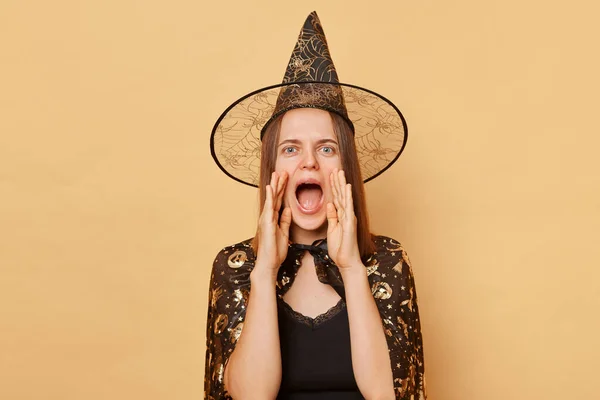 ハロウィーンを祝う魔女のコスチュームとカーニバル円錐形の帽子を身に着けている興奮した若い女性は ベージュの背景に孤立し 口の近くで手で大声で叫び声を上げました — ストック写真
