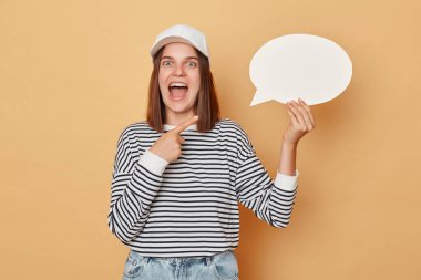 Düşünceli bir posterle reklam yapmak. Heyecanlı kadın çizgili gömlek ve beyzbol şapkası takıyor elinde boş beyaz konuşma balonu bej arka planda tek başına duruyor.
