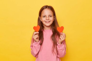 Gülümseyen, iyimser, pembe kazak giyen, sarı arka planda küçük kırmızı kalpler taşıyan, sevgi ve romantik duyguları ifade eden küçük kız..