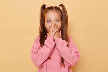 Şok olmuş, at kuyruklu pembe kazaklı küçük bir kız bej arka planda izole edilmiş ağız ve korku dolu gözlerle bakıyor..