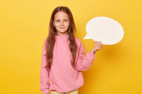 Creative speech bubble for communication. Blank space. Smiling little girl with long hair holding empty speech bubble wearing pink sweatshirt isolated over yellow background
