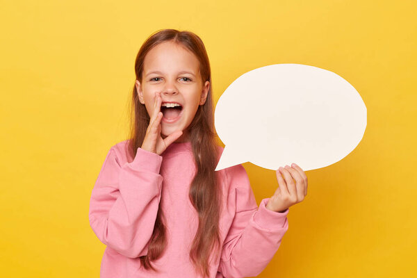 Overjoyed cheerful smiling little girl with long hair holding empty speech bubble wearing pink sweatshirt isolated over yellow background screaming loud making announcement