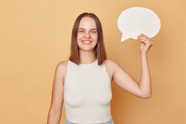 Boş hayal et. Yaratıcı teklif. Boş kafa. Gülümseyen, kahverengi saçlı, günlük kıyafetler giyen, elinde boş baloncuk konuşma bulutu tutan, bej arka planda izole bir şekilde duran genç bir kadın.