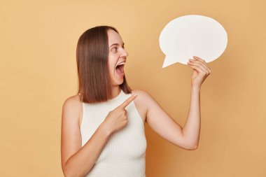 Heyecanlı, kahverengi saçlı, günlük giysiler giymiş genç bir kadın boş baloncuklu konuşma bulutunu işaret ediyor. Düşüncelerini bej arka planda izole bir şekilde sunuyor.