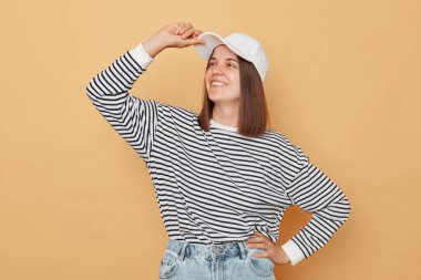 Beyzbol şapkası ve çizgili gömlek giyen kendinden emin bir kadın bej arka planda izole edilmiş gururlu bir yüz ifadesiyle diğer tarafa bakıyor elini kalçasında mutlu bir yüz tutuyor.