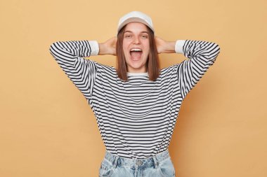 Beyzbol şapkası ve çizgili gömlek giyen aşırı mutlu kadın bej arka planda izole edilmiş neşeli bir yüz ifadesiyle çığlık atıyor.