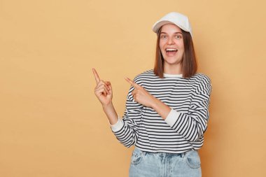 Beyzbol şapkası takan neşeli bir kadın ve bej arka planda izole edilmiş çizgili gömlek reklam alanı için fotokopi alanını işaret ediyor.