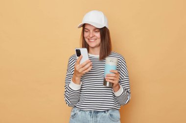 Beyzbol şapkası takan, çizgili gömlek giyen, termos tutan ve bej arka planda izole edilmiş akıllı telefon kullanan neşeli, pozitif kadın sosyal ağları kontrol ediyor.