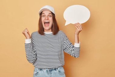 Beyzbol şapkası ve çizgili gömlek giyen aşırı mutlu bir kadın bej arka planda izole edilmiş boş baloncuk konuşma bulutu tutuyor ve sıkılmış yumruk diye bağırıyor.