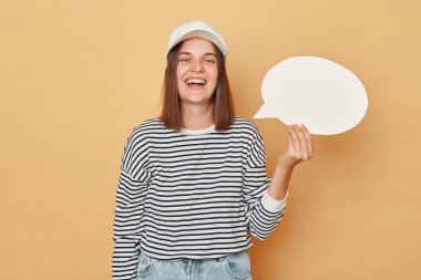 Gülen kadın beyzbol şapkası ve çizgili gömlek giyiyor. Elinde boş balon konuşma bulutu var. Promosyon metni için bej arka plan sunucu alanı üzerinde izole edilmiş.