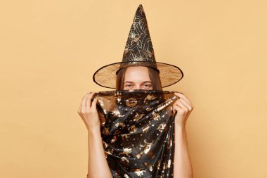 Siyah cadılar bayramı kıyafeti giymiş komik gülümseyen genç kız ve koni bej arka planda izole edilmiş poz veriyor yüzünü gizliyor ve tatil kutlamalarına bakıyor.